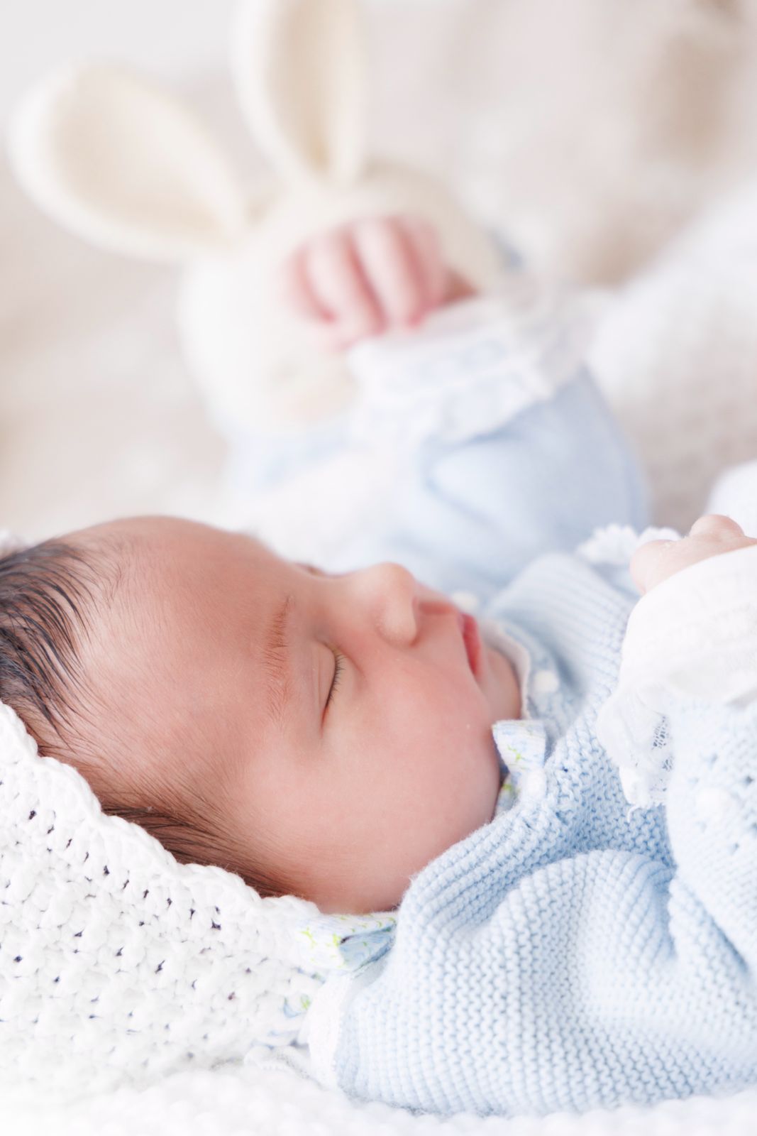 Primer plano lateral de un bebé recién nacido durmiendo plácidamente con ropa de punto azul y un peluche de conejo al fondo.