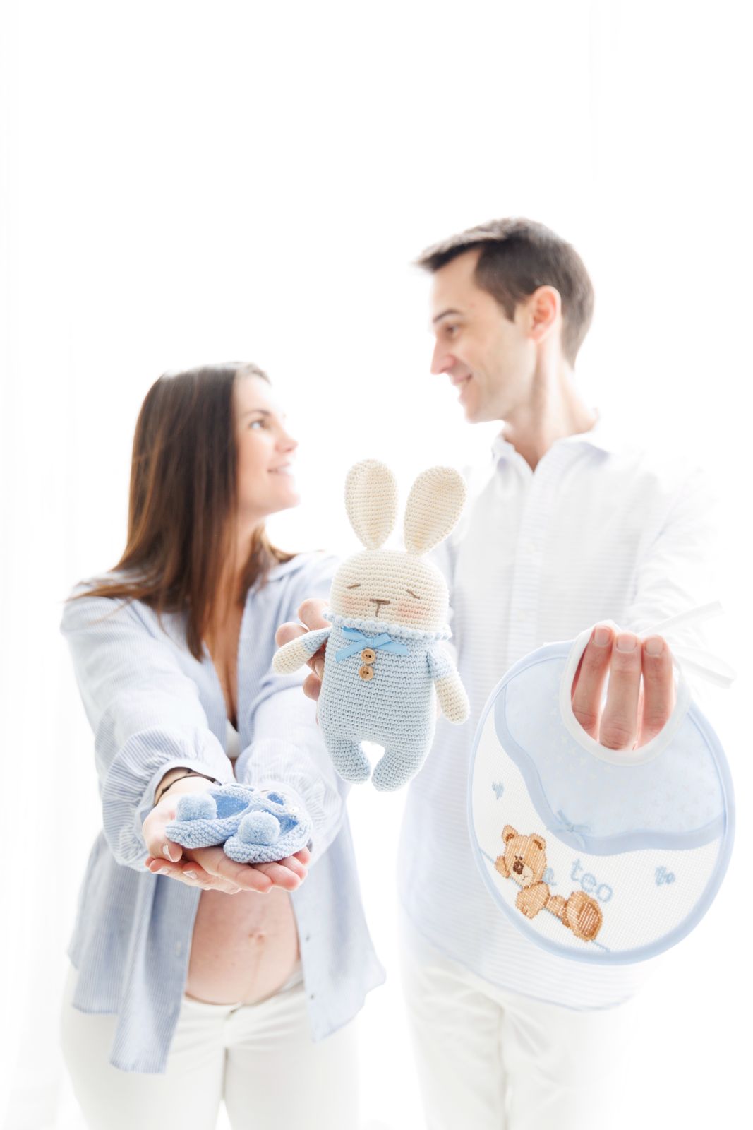 Pareja embarazada desenfocada al fondo mostrando accesorios de bebé en primer plano: patucos, peluche y babero.