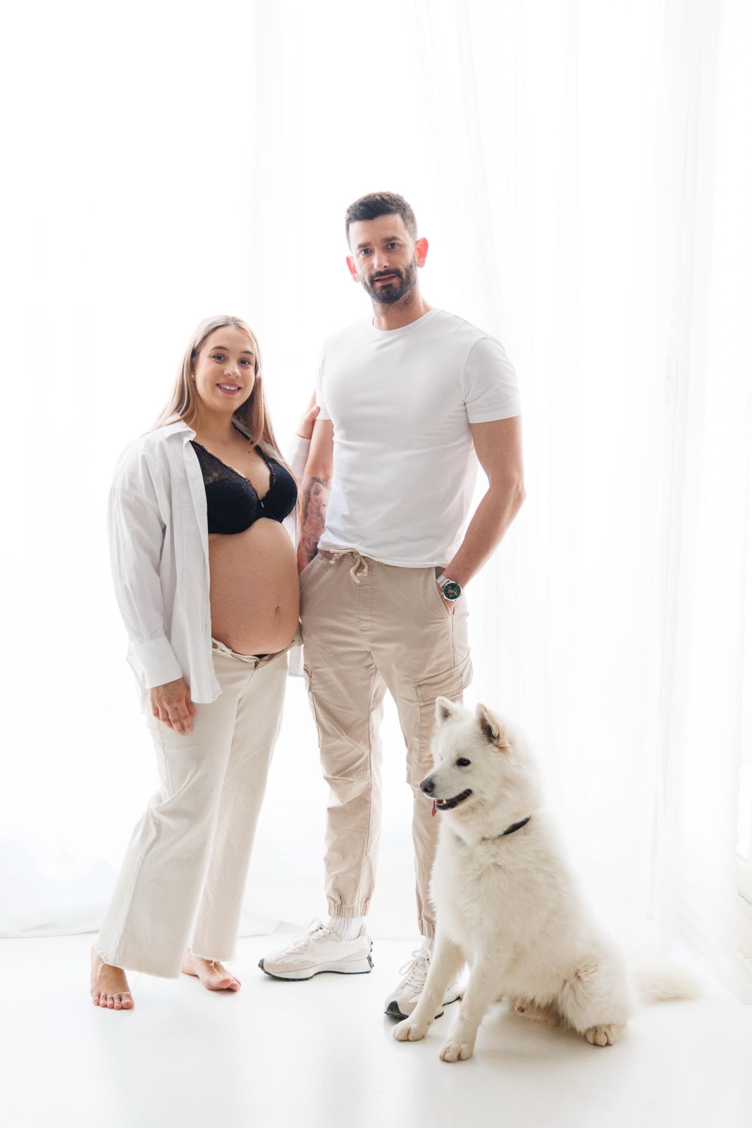 Retrato de estudio de una pareja embarazada posando con su perro blanco sobre un fondo blanco luminoso.
