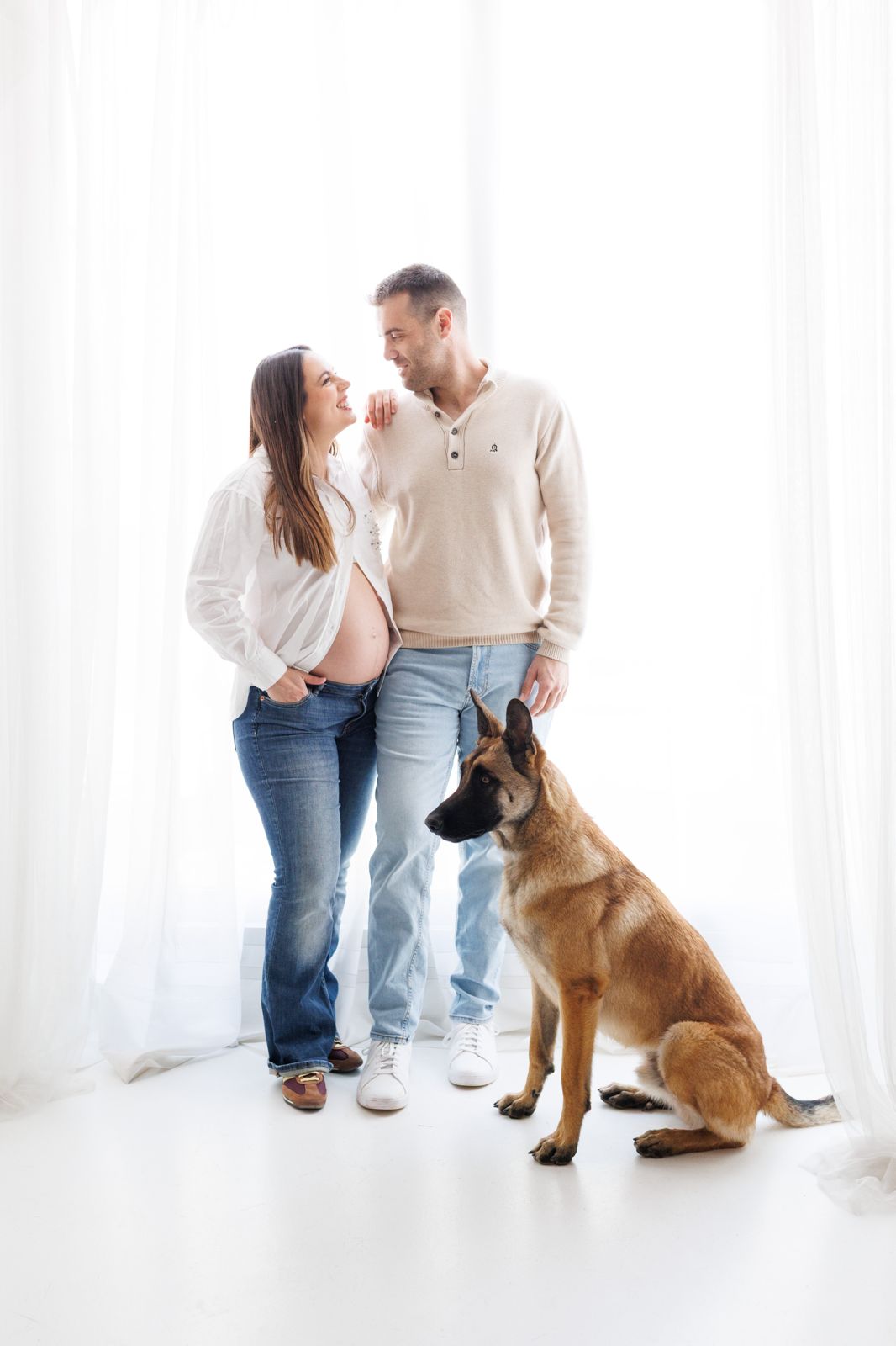 Pareja en espera de un bebé posando alegremente en estudio junto a su perro pastor belga ante un fondo de cortinas blancas.