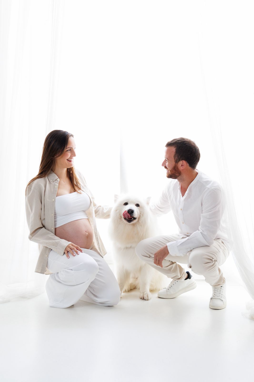 Pareja joven posando en estudio blanco durante embarazo junto a su perro Samoyedo, ambiente luminoso y cálido.