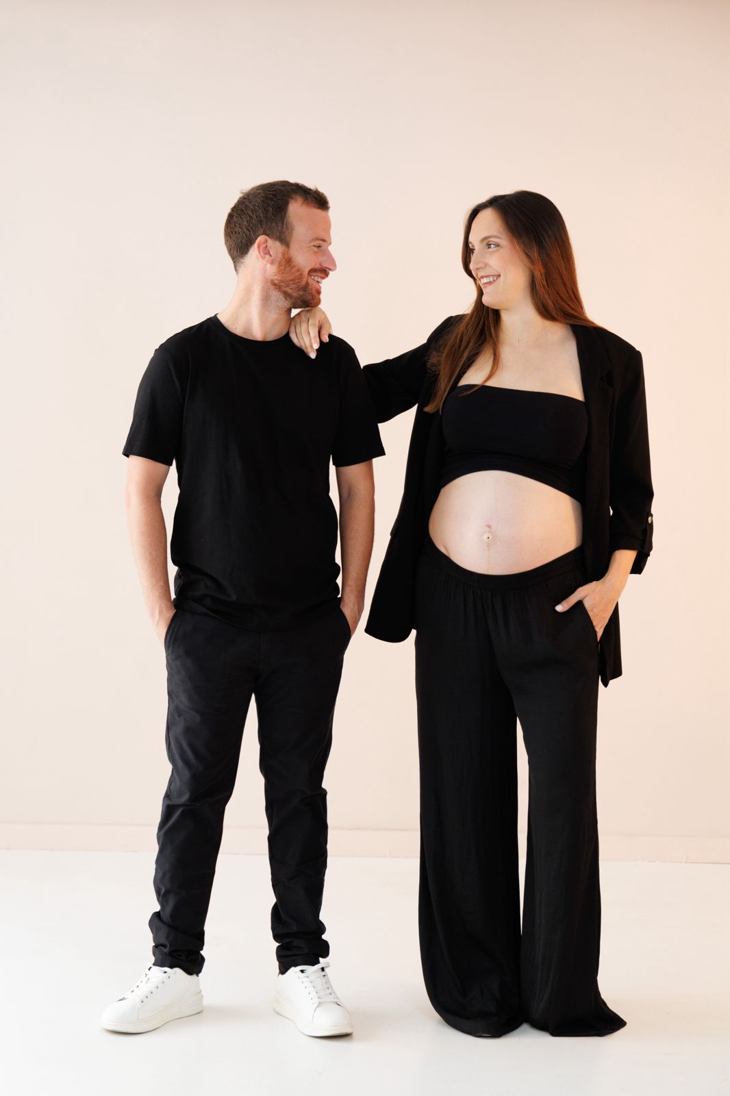 Retrato de estudio de una pareja joven sonriendo, con la mujer embarazada mostrando su vientre, ambos vestidos de negro sobre fondo neutro.