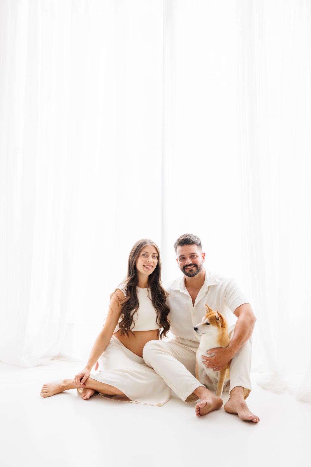 Pareja joven embarazada sentada en el suelo de un estudio blanco junto a su perro Shiba Inu, vistiendo ropa clara en un ambiente luminoso.