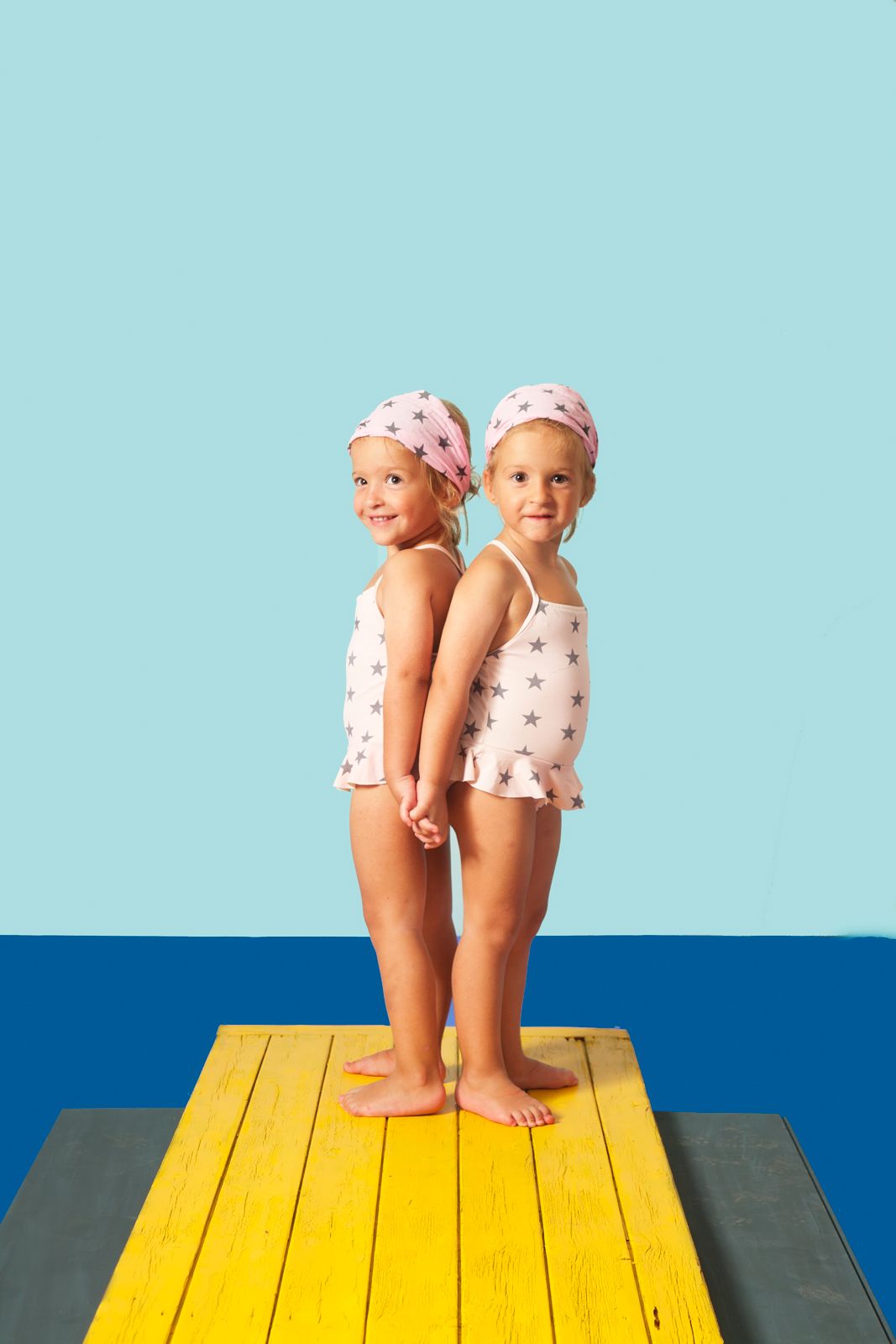 Dos niñas pequeñas posando espalda con espalda con bañadores de estrellas sobre una pasarela amarilla en un estudio fotográfico.