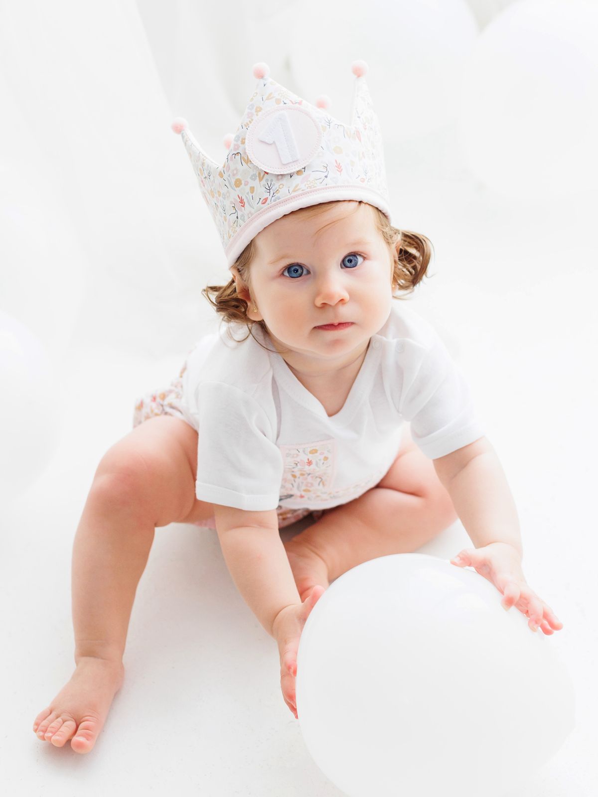 Niña celebrando su primer año con corona de tela y globos blancos en un estudio fotográfico minimalista.