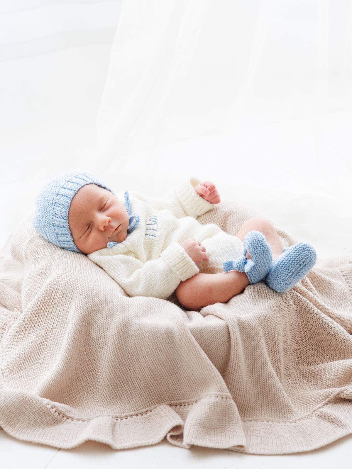 Recién nacido durmiendo plácidamente sobre una manta de punto, vistiendo conjunto blanco con gorro y patucos azules.