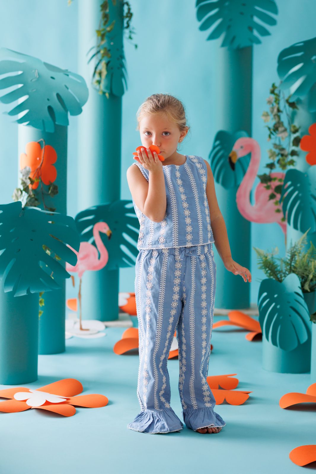 Niña posando en un set fotográfico con temática tropical azul, flamencos rosas y flores de papel.