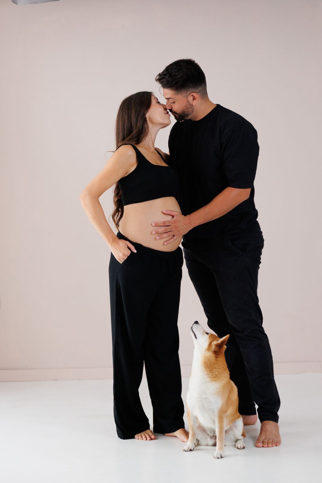 Retrato de estudio de una pareja embarazada besándose junto a su perro sobre un fondo neutro.