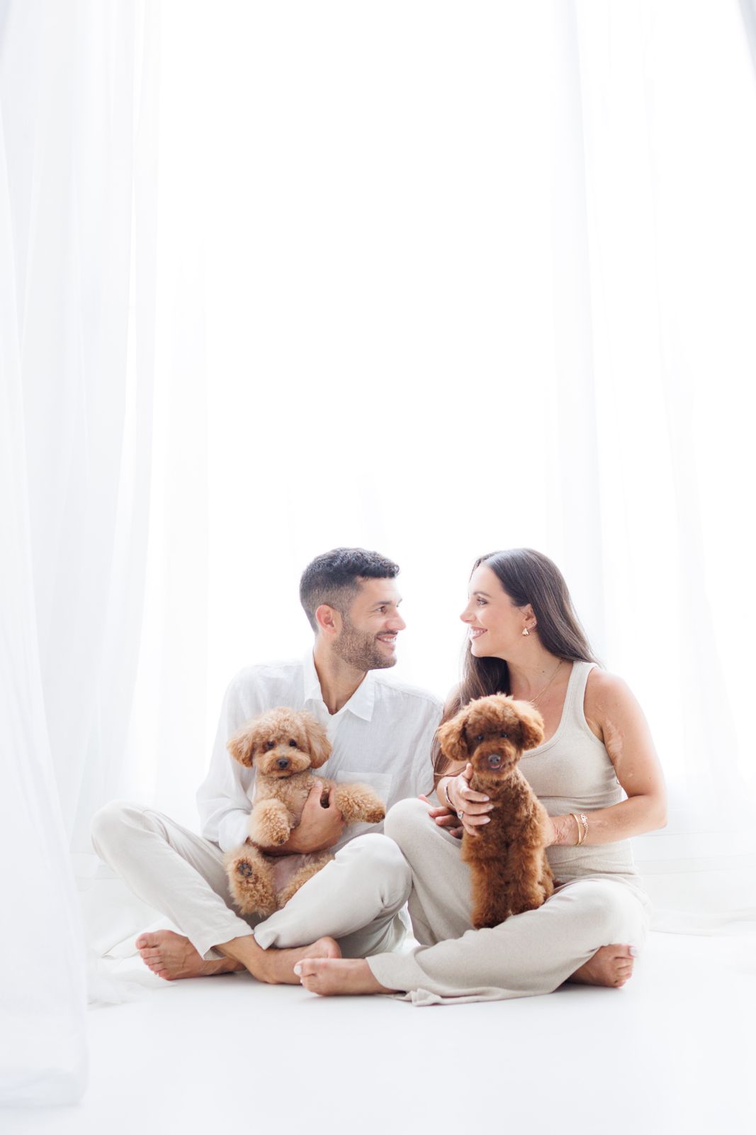 Una pareja joven sentada en el suelo de un estudio luminoso abrazando a sus dos perros caniche toys mientras se miran con ternura.