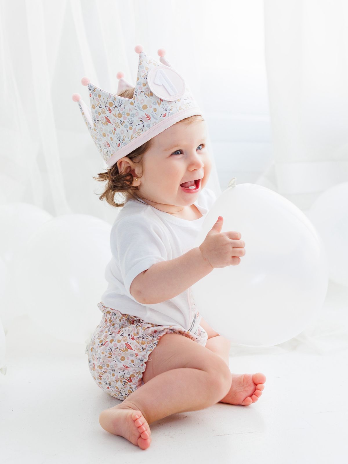 Bebé sonriente de un año con corona de tela y globos blancos en una sesión de fotos de estilo minimalista.