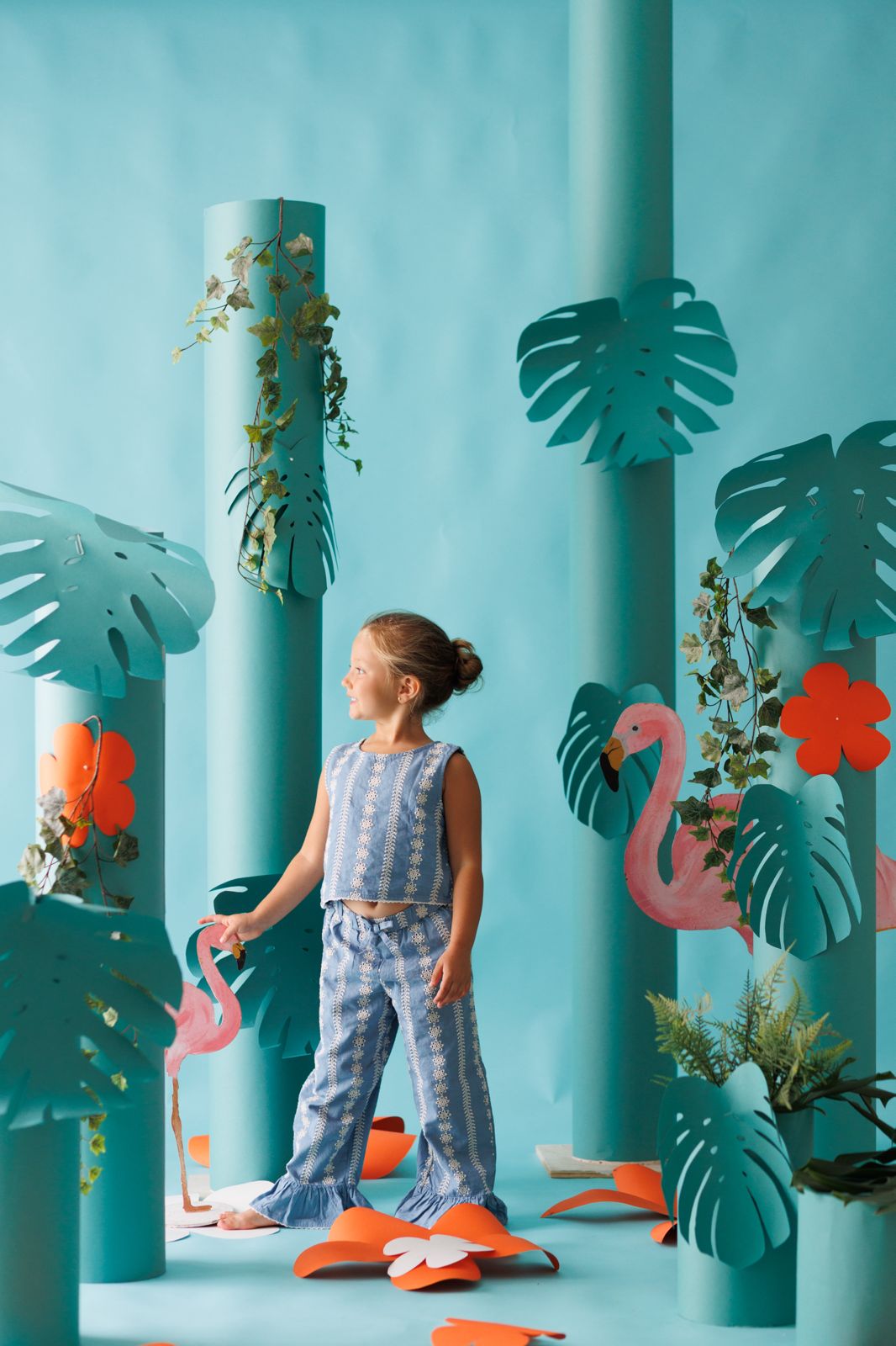 Niña posando en un set fotográfico temático con flamencos rosas, hojas de costilla de Adán y columnas turquesas.