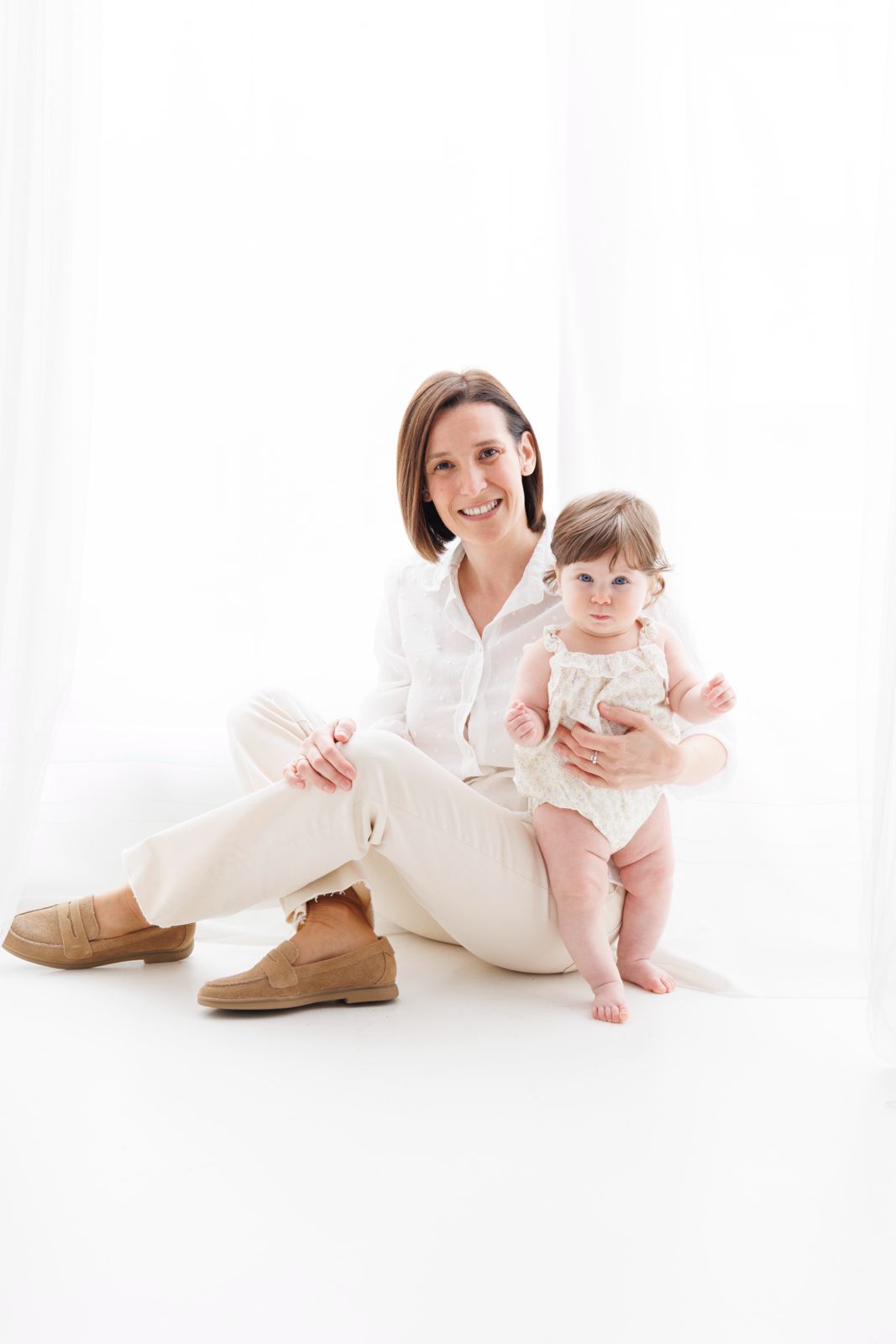 Retrato de estudio en clave alta de una madre sentada en el suelo junto a su bebé, ambas con ropa de tonos claros y fondo blanco.