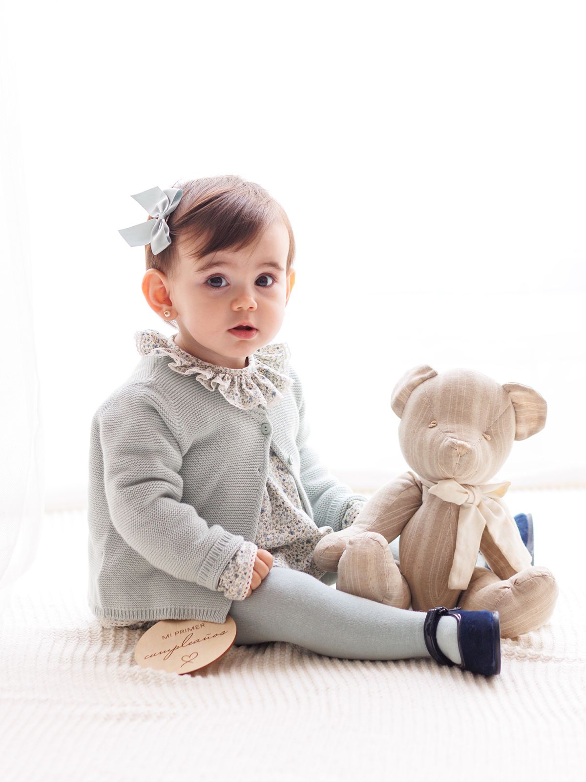 Niña pequeña sentada en un estudio fotográfico junto a un osito de peluche y un disco de madera que indica su primer cumpleaños.